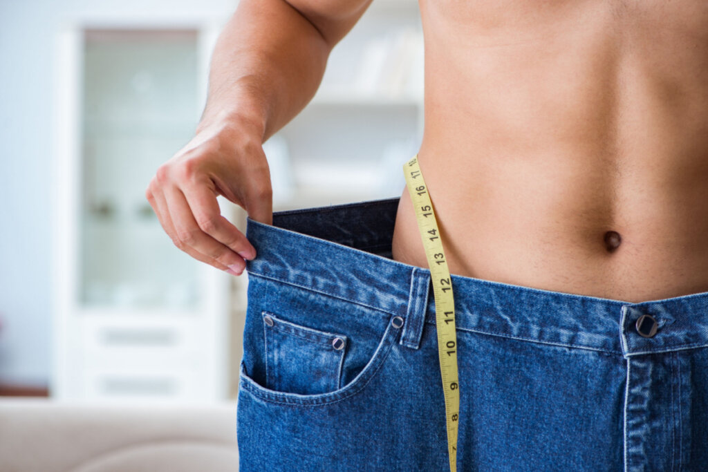 Homem com fita métrica em volta da cintura e mostrando a quantidade de peso que perdeu esticando a calça larga