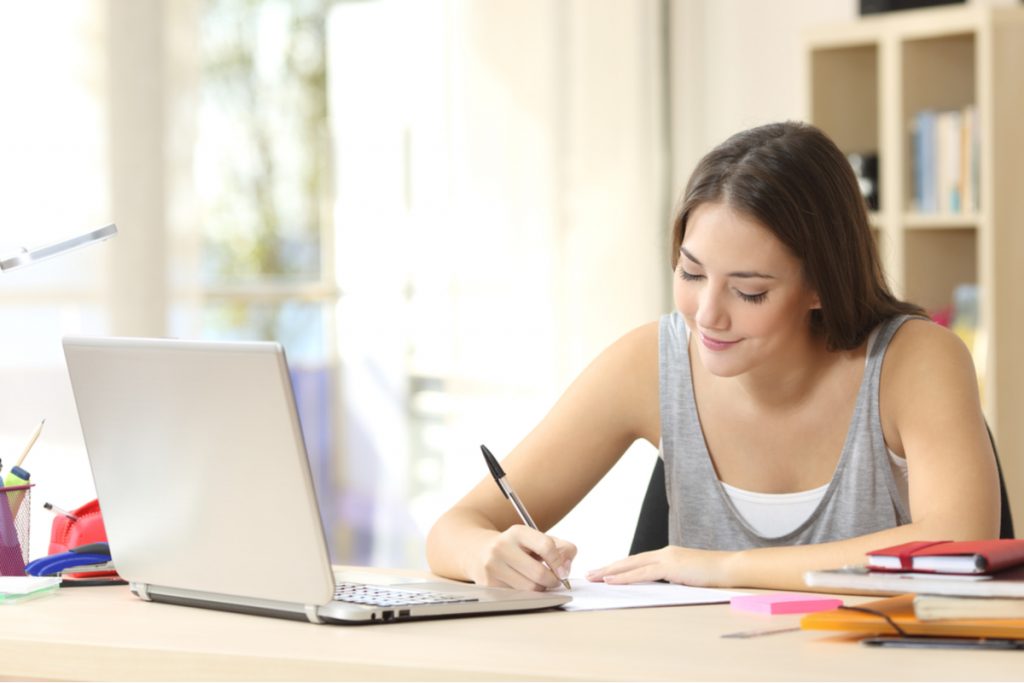 Menina sentada com computador aberto e escrevendo no caderno com uma caneta preta