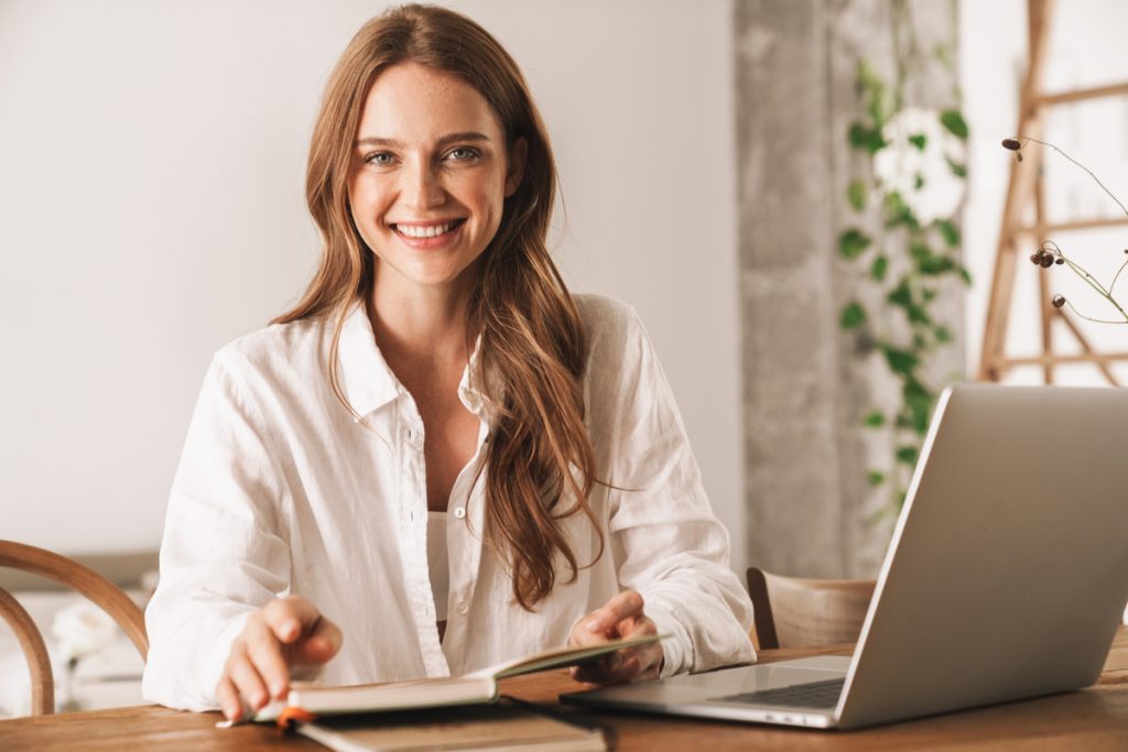 Mulher com semblante alegre, no escritório, usando o laptop e segurando o notebook