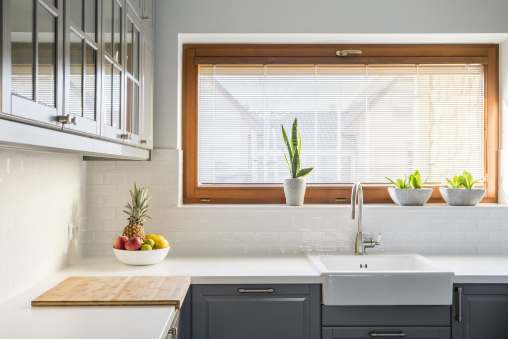 Cozinhe com móveis cinza, bancada branca, janela e vasos de planta