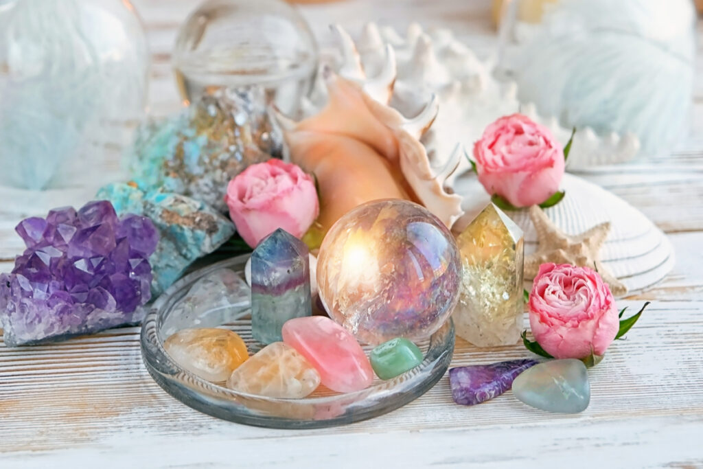 Cristais, flores e conchas coloridos e distribuidas em mesa de madeira branca.