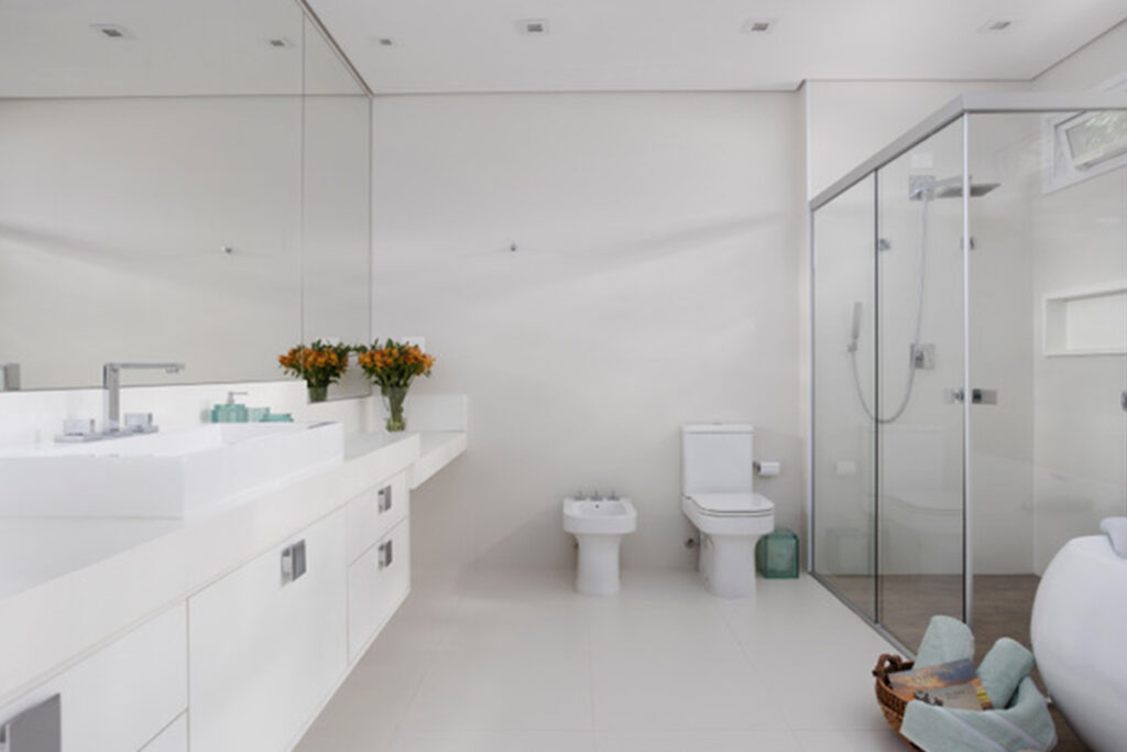 Banheiro com paredes brancas, espelho, móveis brancos e box 
