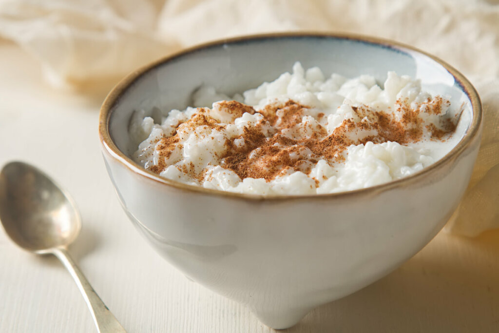 Recipiente branco com arroz-doce polvilhado com canela em pó em cima de uma mesa branca com uma colher do lado 