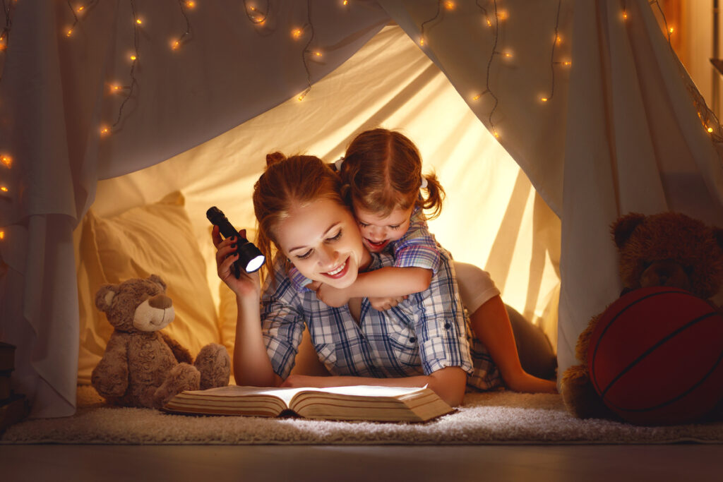 Mulher contando história para criança em barraca improvisada. Com lanterna ligada em cima de livro e ursinho de pelúcia ao lado.