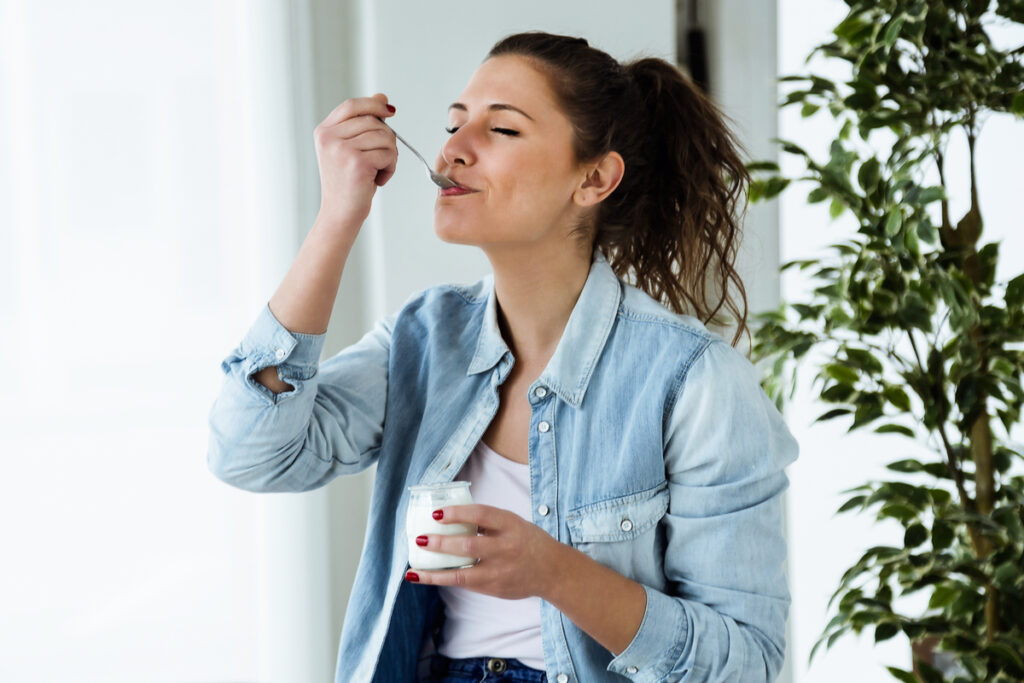Mulher com blusa branca e jaqueta jeans comendo iogurte