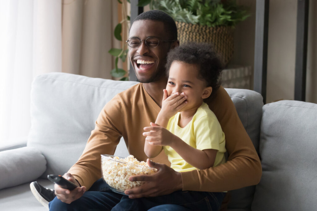 Pai e filho assistindo à televisão sentados em um sofá sorrindo e comendo pipoca