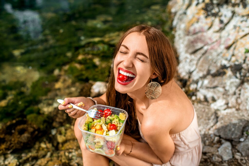 Mulher sorrindo com tomate na boca e segurando um garfo e um recipiente com salada