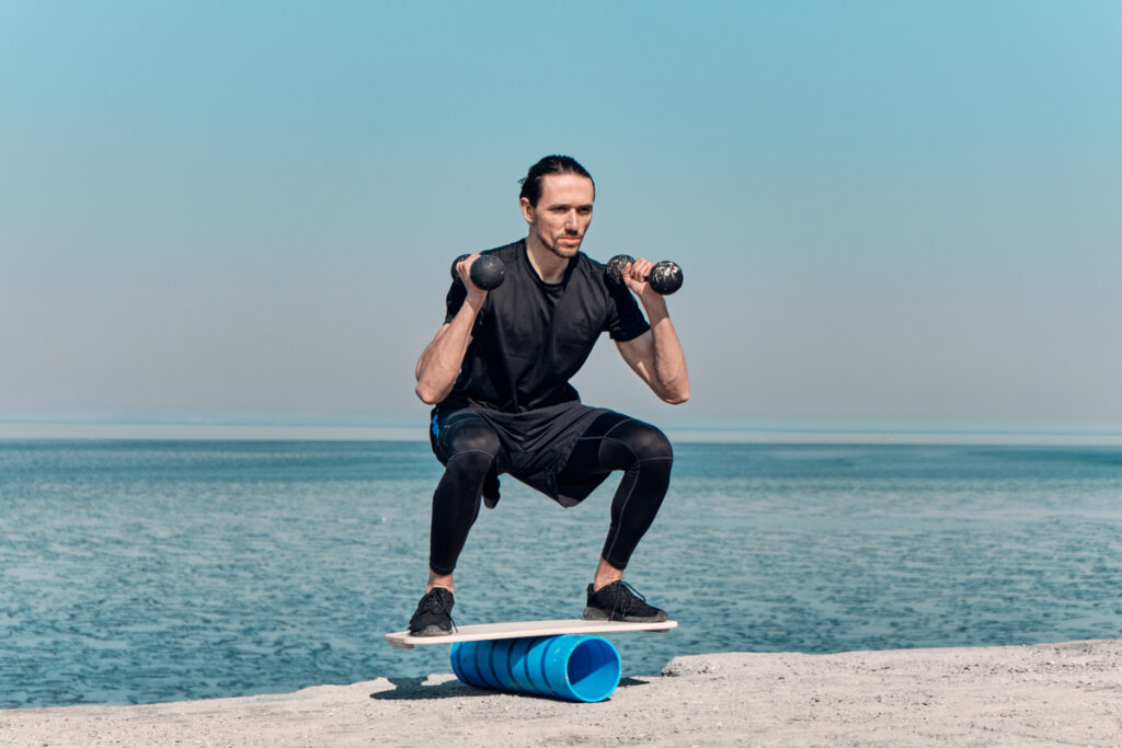 Homem em cima de prancha e agachado com pesos realizando exercício de equílibrio.