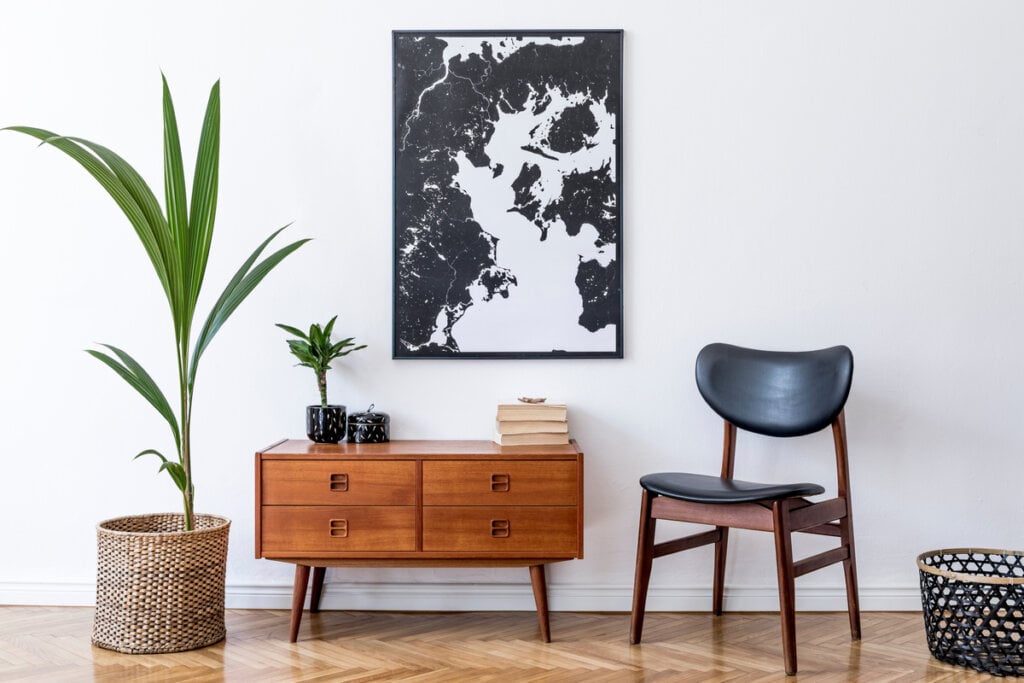 Quadro com imagem abstrata que remete à mármore em preto e branco. Abaixo, uma mesinha de madeira com livros, um vaso e uma planta, uma cadeira e um cesto. 