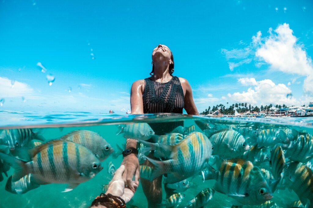 Mulher vestindo um maiô preto dentro do mar fazendo mergulho com peixes 