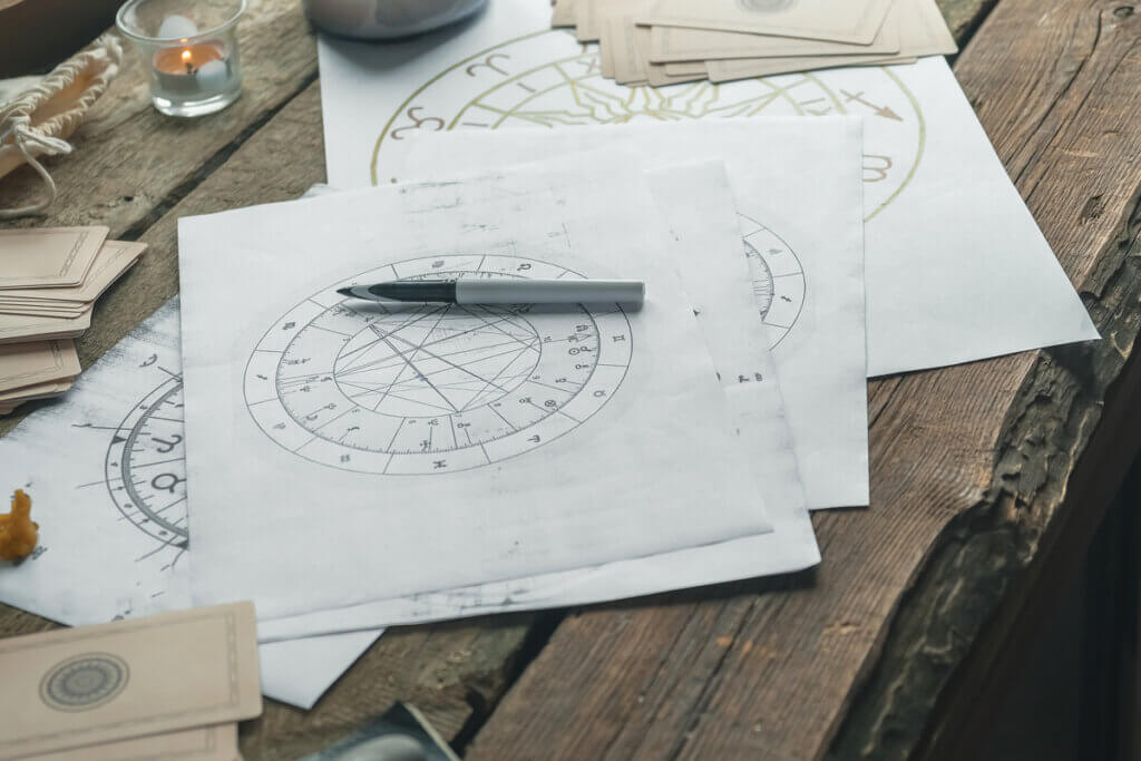 Mesa de madeira com papéis espalhados com desenhos do mapa astral. Na imagem, também há uma caneta, vela e cartas