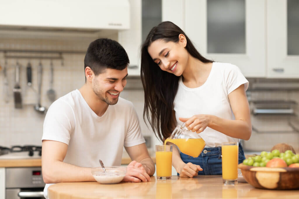Mulher servindo um copo de suco e ao seu lado um homem sorrindo. Na imagem contém uma cesta de fruta e um pote de iogurte