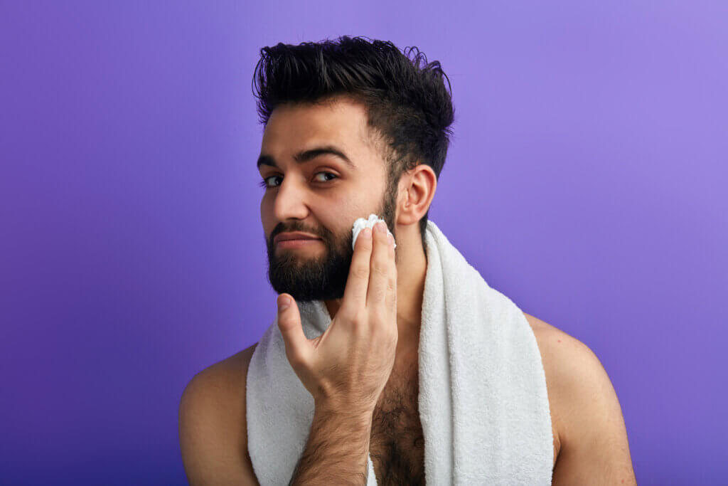 Homem aplicando espuma na barba e com uma toalha enrolada no pescoço no fundo roxo