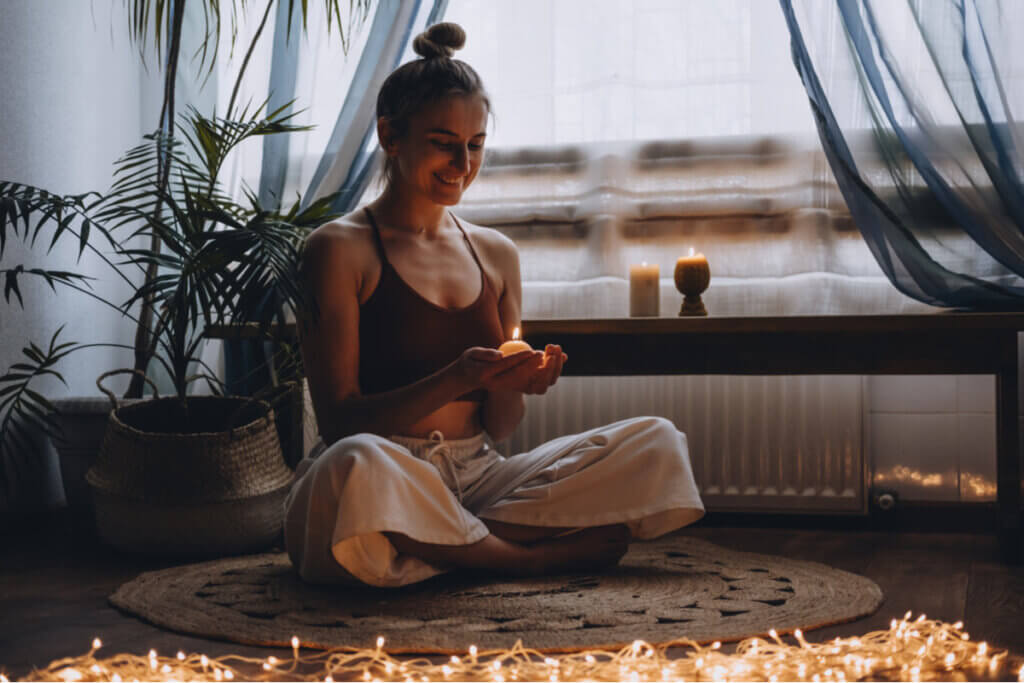 Mulher sentada no chão em posição de meditação, segurando uma vela e rodeada por luzes acesas
