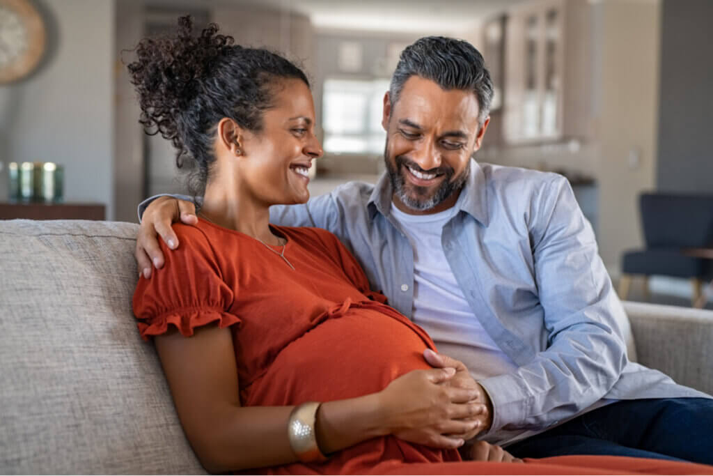 Mulher grávida sentada ao lado de um homem com a mão na barriga dela. Eles estão sorrindo