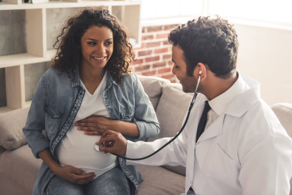 Mulher grávida com a mão na barriga e médico ao lado examinando com estetoscópio