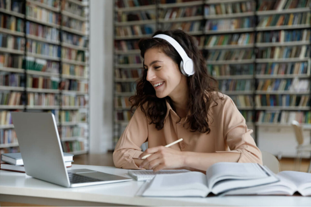 Mulher usando fones de ouvido sentada de frente para o computador estudando e com livros abertos na mesa