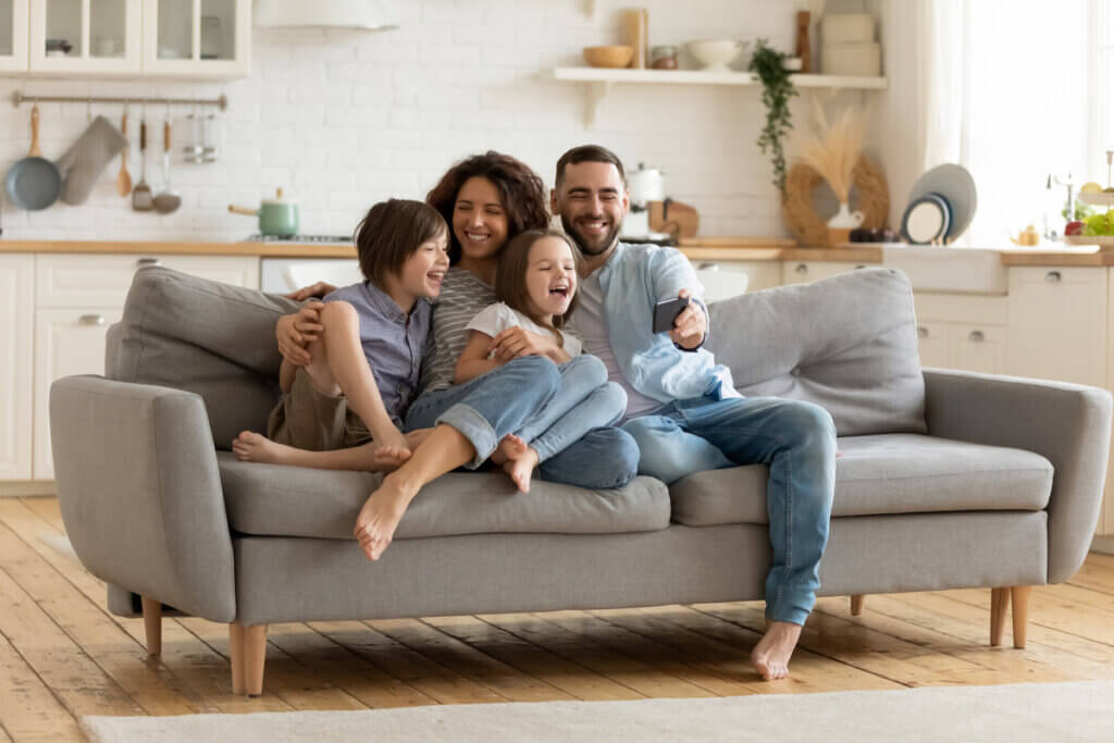 Família feliz sentada no sofá cinza da sala com uma cozinha ao fundo