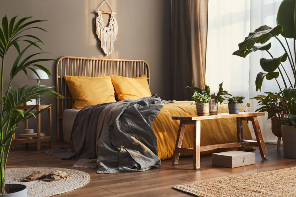 Vista do quarto com roupa de cama amarela, cobertor cinza, banco em madeira e plantas ao redor