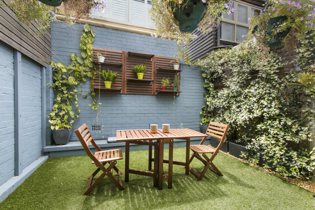 Jardim externo com mesa e cadeira em madeira, parede azul e plantas ao redor