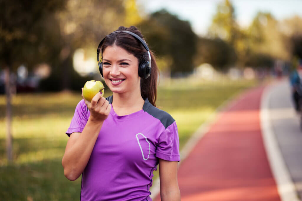 Mulher caminhando ao ar livre com fone de ouvido e segurando uma maçã-verde na mão