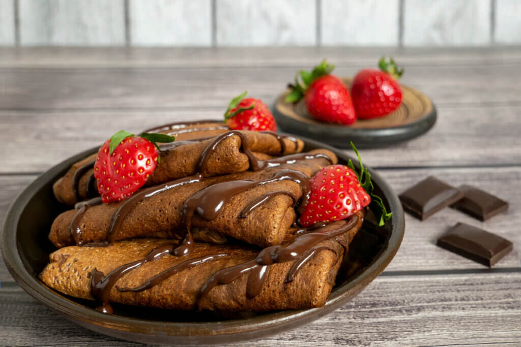 Crepes de chocolate em recipiente marrom, cobertos por calda de chocolate e com morangos 