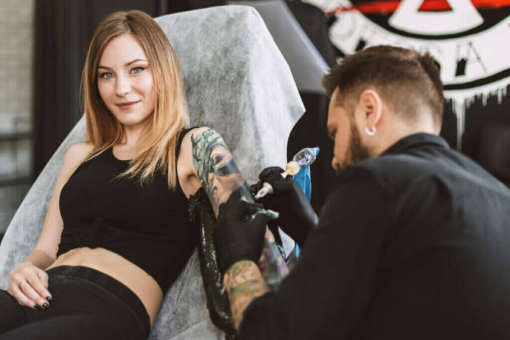 Mulher sorrindo sentada com um tatuador tatuando o seu braço 