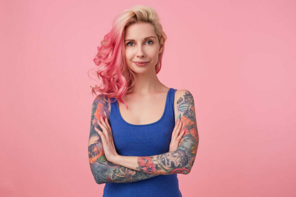 Mulher com cabelo loiro e rosa, tatuada,vestindo blusa azul em frente a um fundo rosa