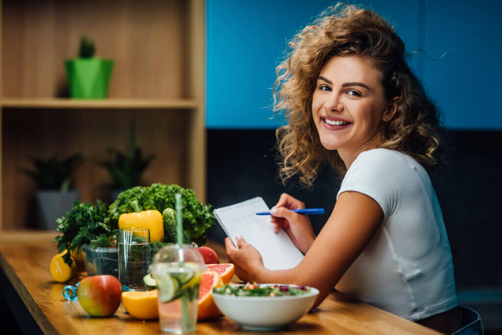 Mulher sentada com caderno e caneta na mão e alimentos saudáveis em cima da mesa