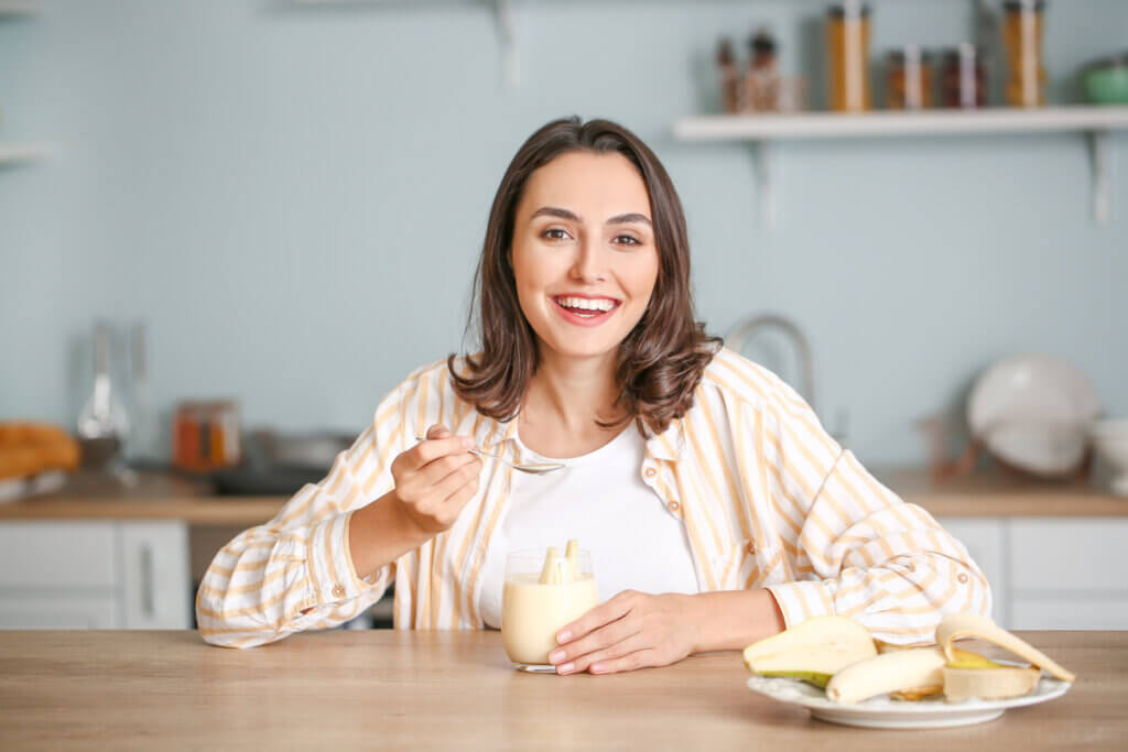 Mulher sentada na cozinha comendo iogurte e um prato de banana do lado