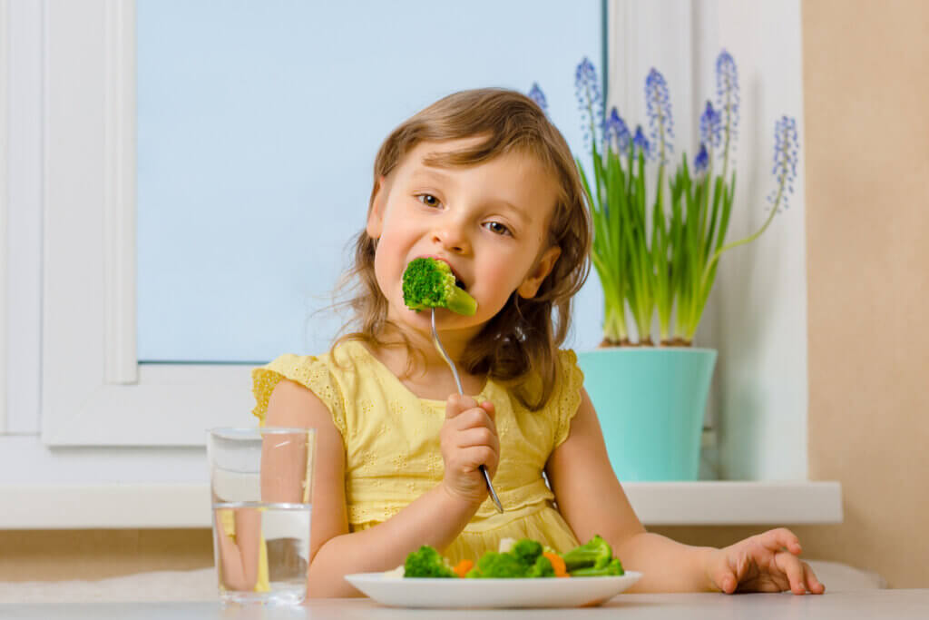 Criança segurando um garfo com um pedaço de brócolis. Na mesa possui um prato com salada e um copo de água