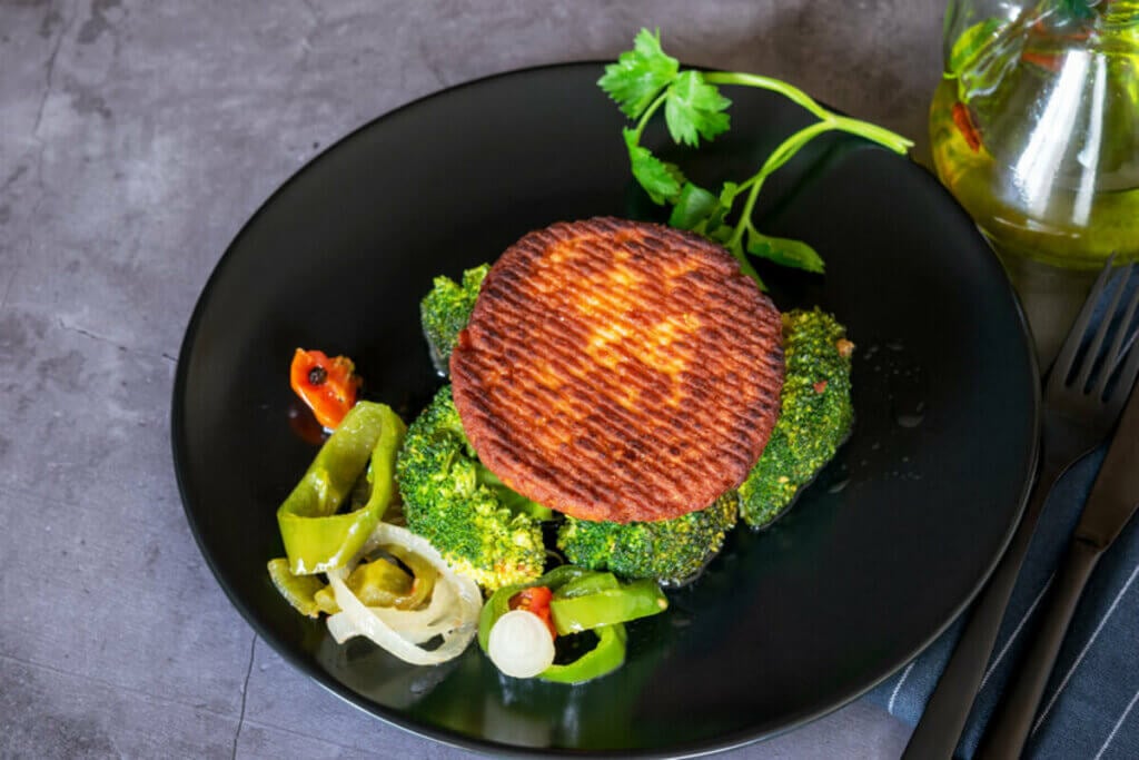 Hambúrguer de salmão em cima de um prato preto com brócolis, cebola, tomate e pimentão