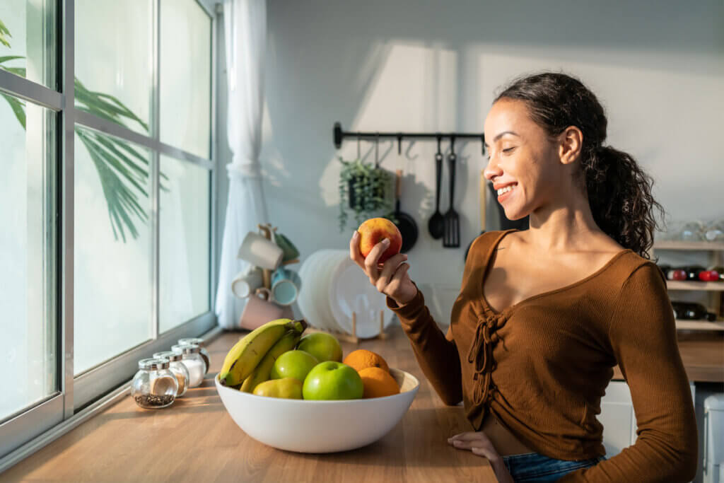 Mulher com fruta na mão. Na mesa, há um bowl com diversas frutas