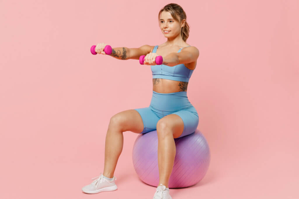 Mulher com roupa de academia, sentada em uma bola levantando peso