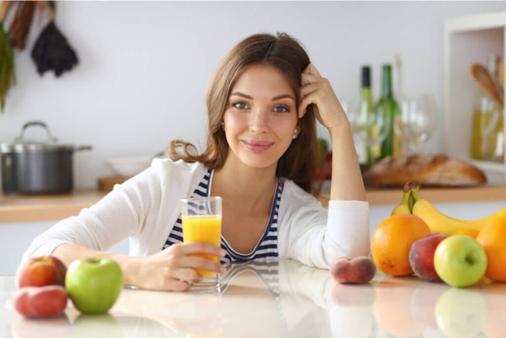 Mulher sentada segurando um copo de suco e frutas (banana, laranja, maçã) em cima da mesa