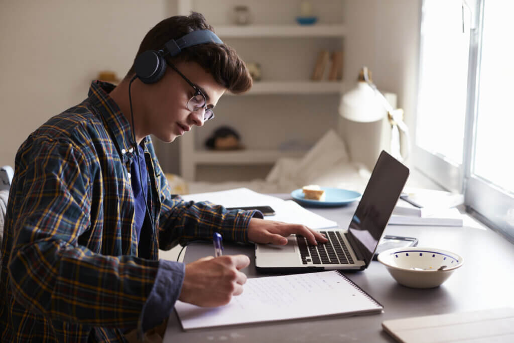 Menino usando fones de ouvido sentado com computador aberto e caneta na mão escrevendo