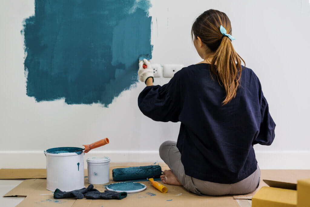 Mulher sentada no chão pintando a parede com pincel. Ao lado tem um balde com tinta e um rolo de pintar