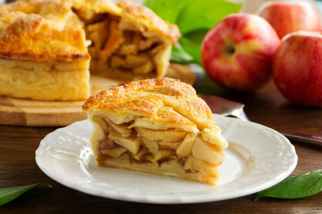 Pedaço de torta de maçã dentro do prato branco, torta inteira e maçãs ao fundo