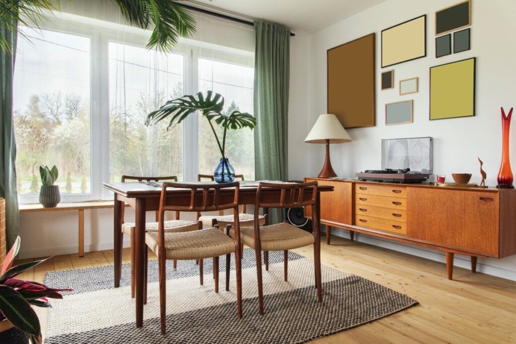 Sala de jantar com estilo tropical; mesa e cadeiras de madeira rústica, parede decorada com quadros geométricos, cortinas verdes e plantas