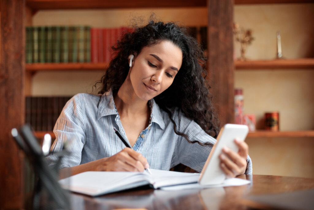Mulher sentada estudando com caderno aberto em cima da mesa, segurando uma lapiseira em uma mão e um celular na outra