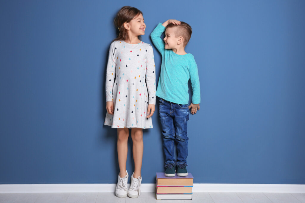 Menino e menina comparando altura em parede azul. Menino é menor e está em cima de pilha de livros