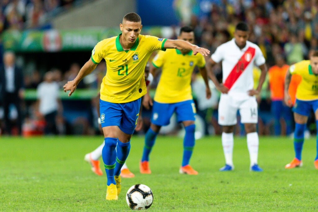 Richarlison batendo falta usando a camisa do Brasil e outros jogadores ao fundo