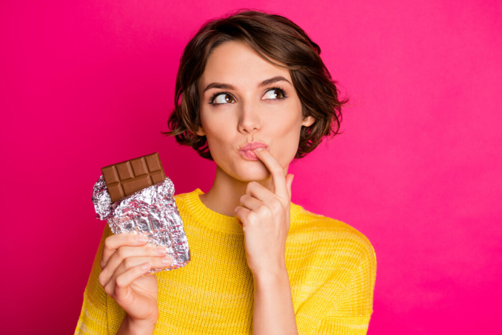 Mulher segurando barra de chocolate com dedo na boca e olhando para o lado