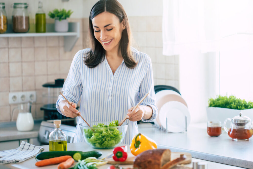 Mulher sorrindo e preparando uma salada e alimentos saudáveis em cima da bancada da cozinha