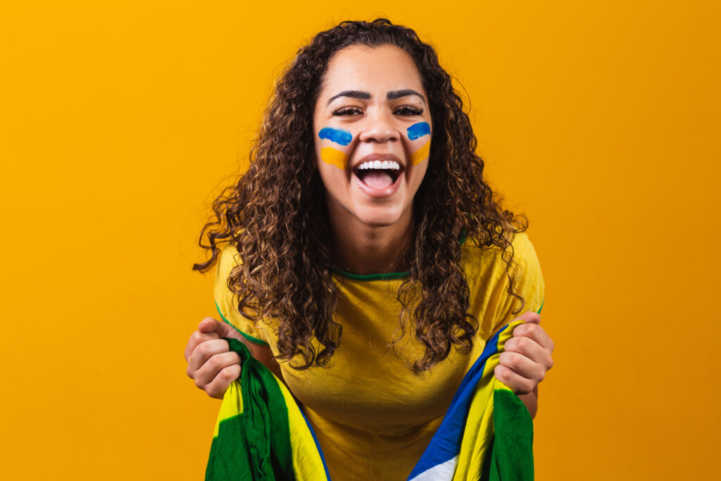 Mulher com o rosto pintado segurando bandeira do Brasil e sorrindo