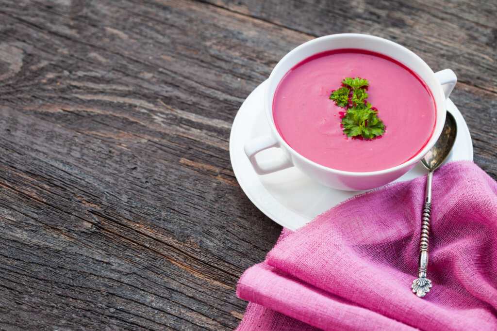 Sopa rosa de beterraba em recipiente e prato branco com pano rosa e colher de metal ao lado