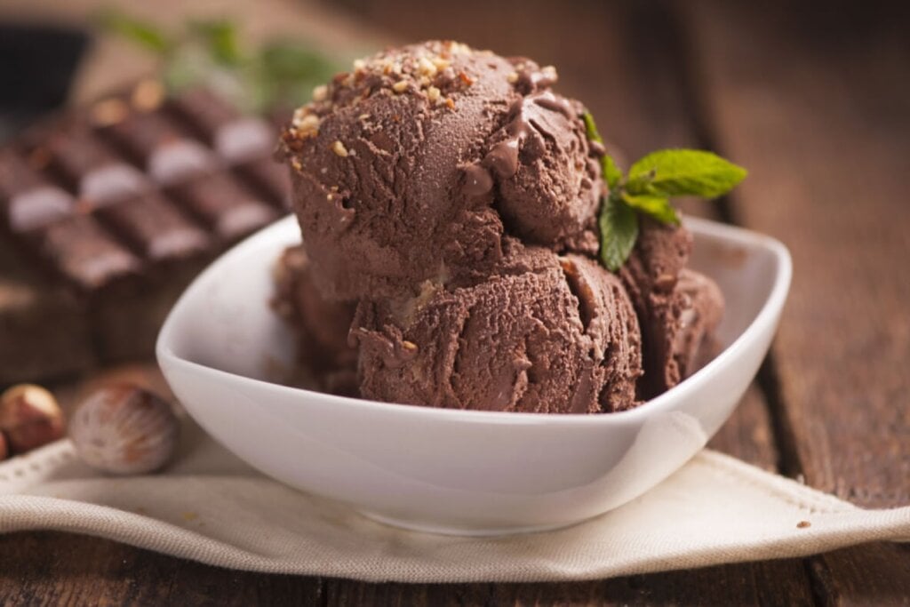 Recipiente branco com duas bolas de sorvete de chocolate decorado com avelãs trituradas e folhas de manjericão