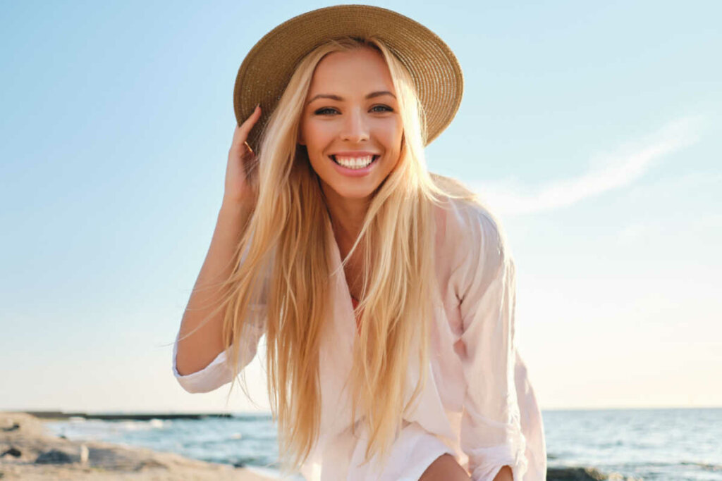 Mulher sorrindo, usando chapéu e praia ao fundo