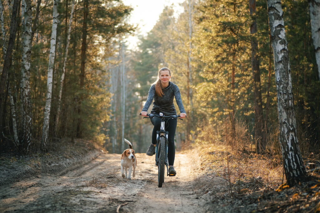 Mulher pedalando e cachorro andando ao lado em meio a floresta