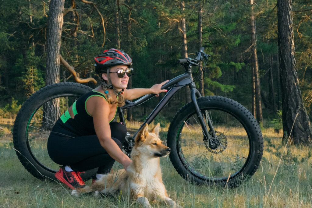 Mulher abaixada segurando bicicleta, com mão em cachorro e olhando para o horizonte 